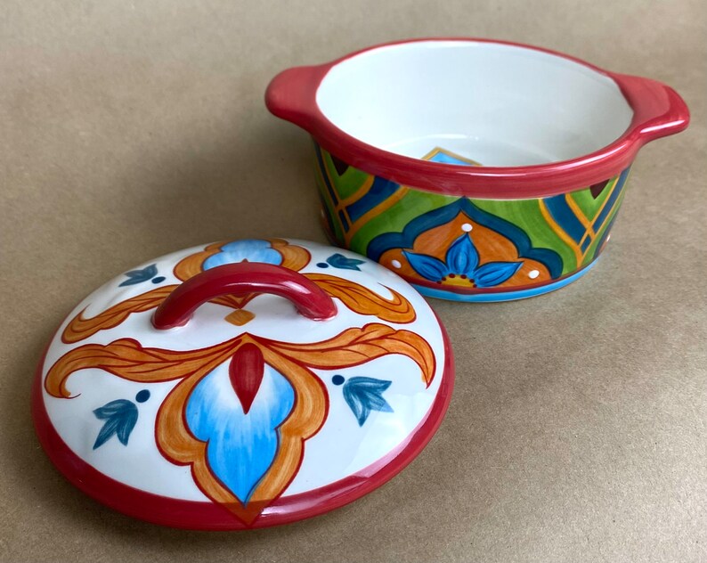 May include: A white ceramic bowl with a red rim and a colorful floral design. The bowl has a matching lid with a red handle.