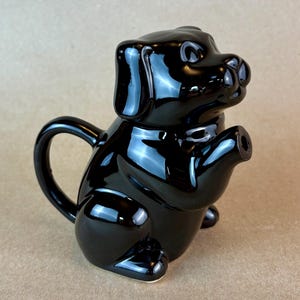 May include: A glossy black ceramic teapot shaped like a dog. The teapot has a curved handle, a spout, and a lid that is part of the dog's head. The dog is sitting upright, with its paws in front.