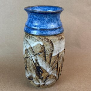 May include: A ceramic vase featuring a flared rim with a vibrant blue glaze. The lower body displays a textured, earthy brown surface with deep, carved grooves and irregular white glaze accents that create a layered, multi-dimensional appearance.