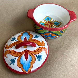 May include: A white ceramic dish with a red rim and a colorful floral design. The dish has a matching lid with a red handle.