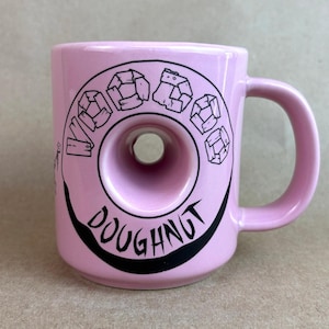 May include: A light pink ceramic mug featuring a central circular cutout that mimics the shape of a doughnut. The mug displays black graphic text that reads Voodoo Doughnut, with the word Voodoo formed by blocky letters and Doughnut below.