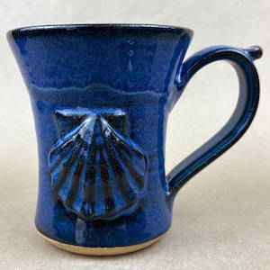 May include: A handmade ceramic mug in a deep blue glaze. The mug features a sculpted seashell design and a curved handle. The interior and base are unglazed, revealing the natural clay color. The mug is suitable for hot or cold beverages.