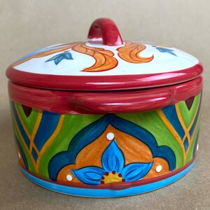 May include: A round ceramic casserole dish with a lid. The dish has a colorful, hand-painted design with blue, green, orange, and yellow accents. The lid has a red handle.