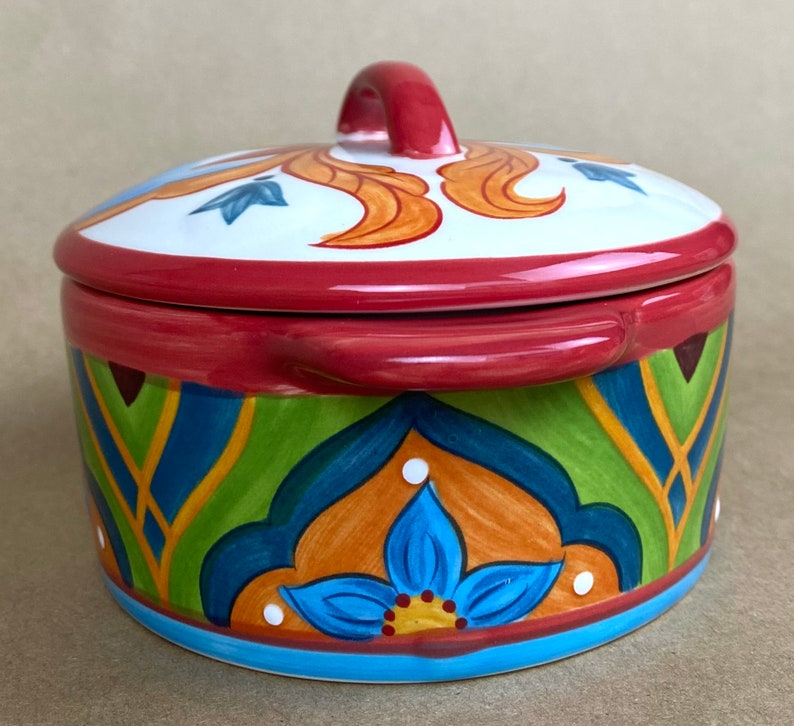 May include: A round ceramic casserole dish with a lid. The dish has a colorful, hand-painted design with blue, green, orange, and yellow accents. The lid has a red handle.