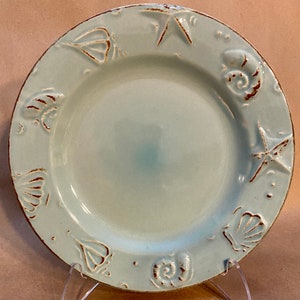 May include: A round, light blue ceramic plate with a brown rim and embossed seashell and starfish designs. The plate has a slightly distressed look, suggesting a coastal or nautical theme. The plate is displayed on a clear stand.