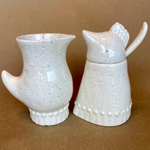 May include: A white ceramic creamer and sugar set shaped like chickens. The creamer has a handle shaped like a chicken wing. The sugar dispenser has a lid shaped like a chicken head. Both pieces have a speckled white glaze.