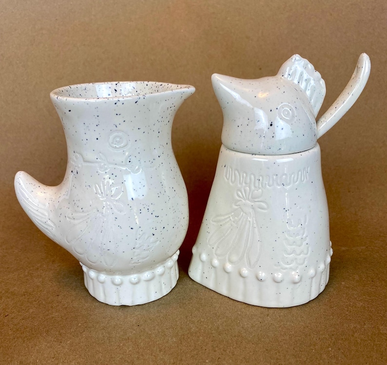 May include: A white ceramic creamer and sugar set shaped like chickens. The creamer has a handle shaped like a chicken wing. The sugar dispenser has a lid shaped like a chicken head. Both pieces have a speckled white glaze.