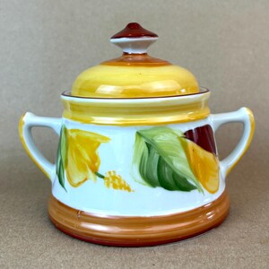 May include: A white ceramic sugar bowl with two handles and a matching lid. The bowl features hand-painted yellow and green floral motifs, with a deep red and orange band around the base and a matching red and orange knob on the lid.