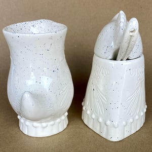 May include: A white ceramic sugar and creamer set shaped like owls. The creamer has a speckled glaze and a small tail. The sugar bowl has a lid shaped like an owl's head and a speckled glaze.