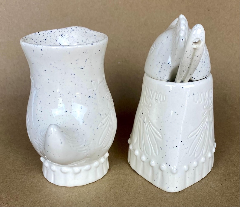 May include: A white ceramic sugar and creamer set shaped like owls. The creamer has a speckled glaze and a small tail. The sugar bowl has a lid shaped like an owl's head and a speckled glaze.