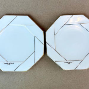 May include: Two white octagonal plates with gold trim and geometric black lines. The plates have the brand name "GIVENCHY PARIS" printed at the bottom. The plates are likely for decorative or serving purposes.