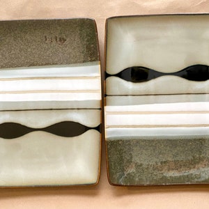 May include: Two square ceramic plates with a distinctive design. Each plate features a dark olive green top section, horizontal stripes in white, light grey, and beige, and a wavy black band. The plates have a brown border.