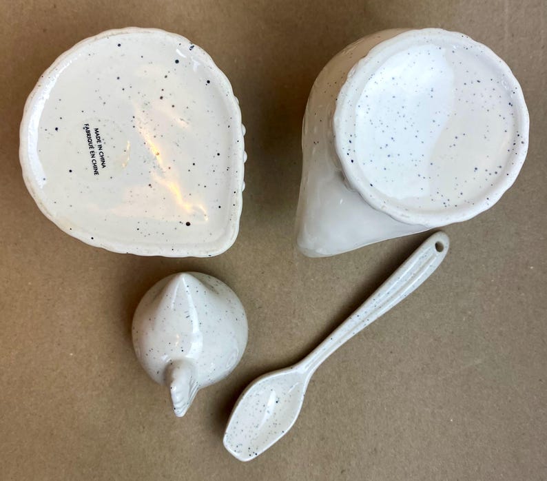 May include: White ceramic chicken-shaped sugar bowl and creamer set with a speckled finish. The set includes a small spoon. The bottom of the sugar bowl reads "Made in China, Fabriqu&eacute; en Chine".