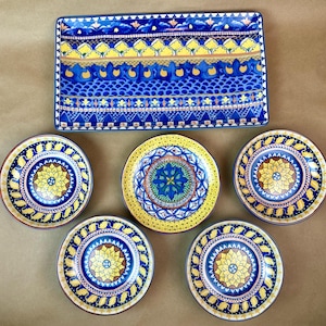 May include: A set of decorative ceramic tableware featuring a rectangular serving platter and five round plates. The blue and yellow plates have intricate floral and geometric patterns. The serving platter has a similar design.