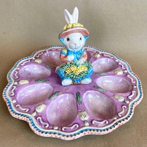May include: A pink ceramic egg holder with a bunny figurine on top. The bunny is holding a bouquet of yellow flowers and is wearing a blue and white dress. The egg holder has six individual egg cups.