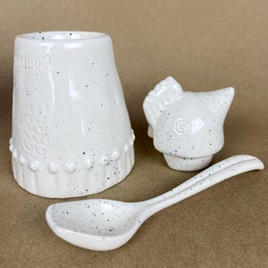 May include: White ceramic sugar dispenser with a chicken-shaped lid and a matching spoon. The dispenser has a speckled white glaze with blue flecks.