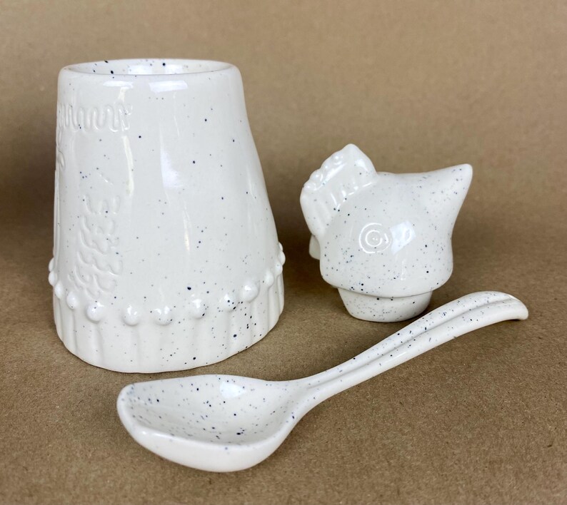 May include: White ceramic sugar dispenser with a chicken-shaped lid and a matching spoon. The dispenser has a speckled white glaze with blue flecks.