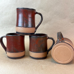 May include: Four handmade ceramic mugs. The mugs are a deep red with black handles and rims. The base of each mug has a tan band with a textured design. One mug is lying on its side, showing the bottom.