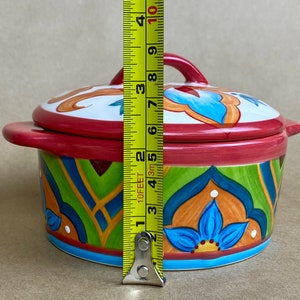 May include: A colorful ceramic casserole dish with a lid. The dish has a red base with green, blue, and orange geometric patterns. The lid has a white base with blue and orange floral designs. The dish measures 6 inches in diameter.