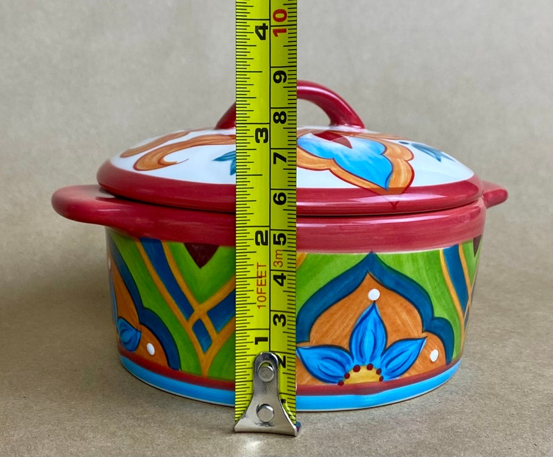 May include: A colorful ceramic casserole dish with a lid. The dish has a red base with green, blue, and orange geometric patterns. The lid has a white base with blue and orange floral designs. The dish measures 6 inches in diameter.