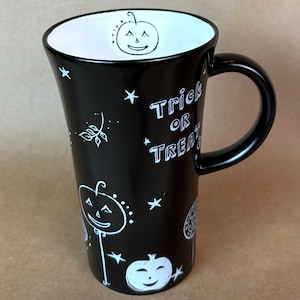 May include: Black ceramic Halloween mug with a white interior and handle. The mug features white illustrations of jack-o'-lanterns, stars, and the words "Trick or Treat". The mug has a tall, tapered shape.