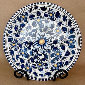 May include: A round ceramic plate featuring a hand-painted floral pattern in shades of dark blue and light blue with yellow centers. The design includes swirling leaves and a central flower, finished with a striped border along the outer edge.