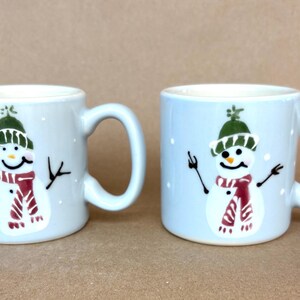 May include: Two light blue ceramic mugs, each featuring a hand-painted snowman wearing a green hat and a red striped scarf. The snowmen have black stick arms, orange carrot noses, and are surrounded by small white dots representing falling snow.