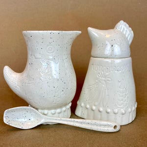 May include: White ceramic creamer and sugar set shaped like chickens with a speckled finish. The creamer has a handle shaped like a chicken wing. The sugar bowl has a lid shaped like a chicken head. A matching speckled spoon is included.