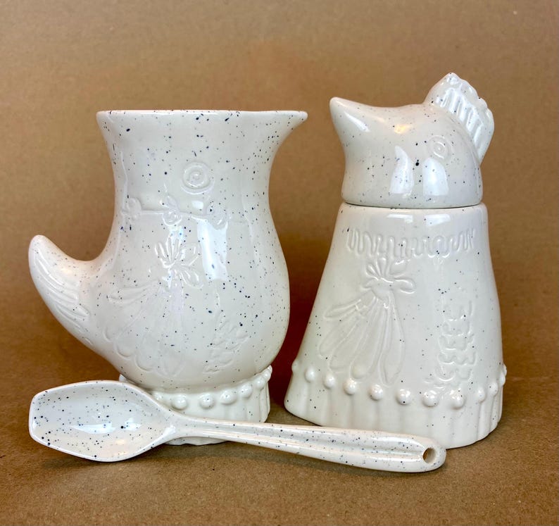 May include: White ceramic creamer and sugar set shaped like chickens with a speckled finish. The creamer has a handle shaped like a chicken wing. The sugar bowl has a lid shaped like a chicken head. A matching speckled spoon is included.