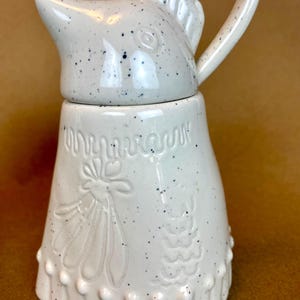 May include: White ceramic jug with a bird-shaped lid and a speckled glaze. The jug has a floral design and a scalloped edge.