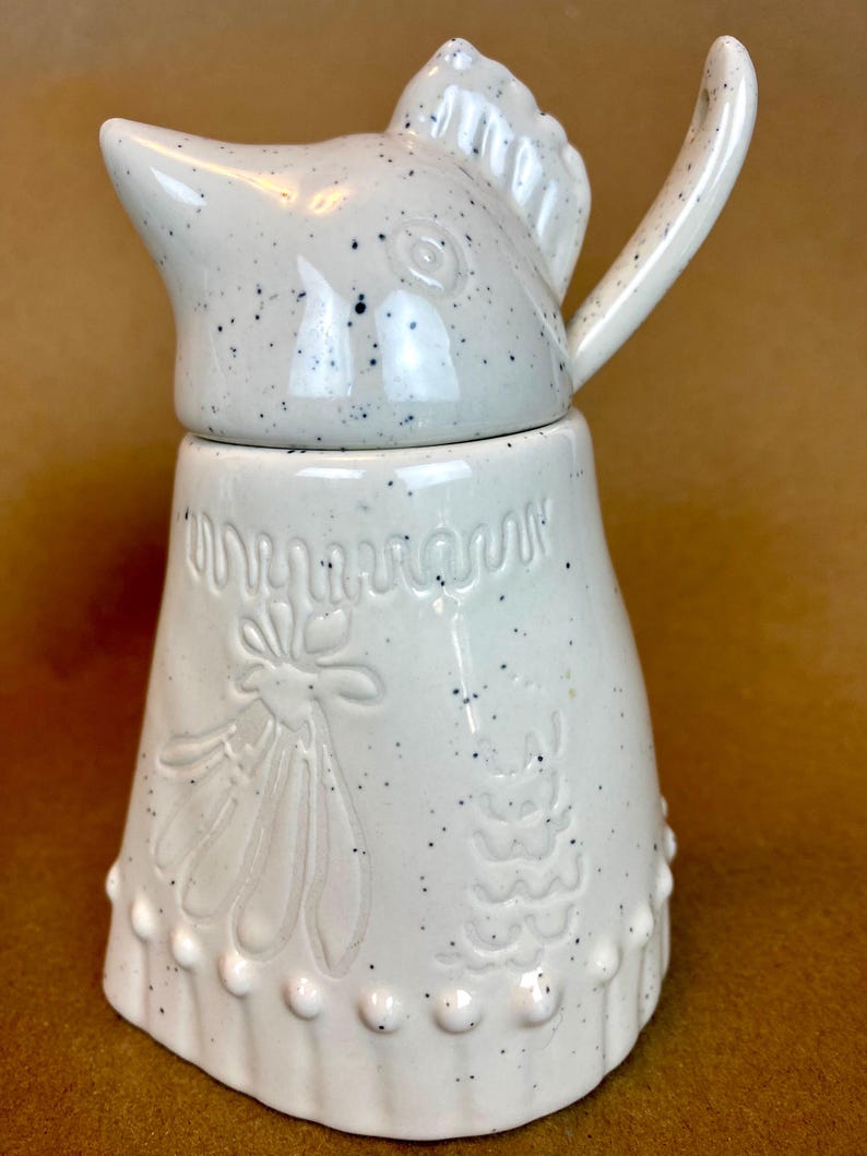 May include: White ceramic jug with a bird-shaped lid and a speckled glaze. The jug has a floral design and a scalloped edge.