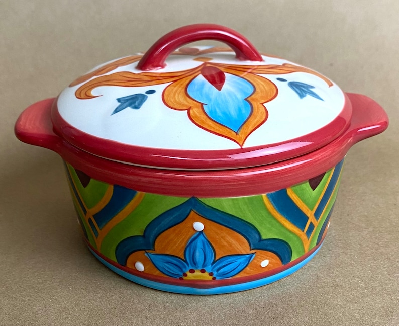 May include: A colorful ceramic casserole dish with a lid. The dish has a red rim and a blue, green, orange, and yellow floral pattern. The lid has a red handle and a similar floral pattern.