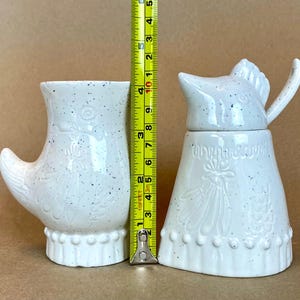 May include: Two white ceramic kitchenware items with a speckled glaze. The item on the left is a pitcher shaped like a boot with a white floral design. The item on the right is a sugar dispenser shaped like a bird with a white floral design.
