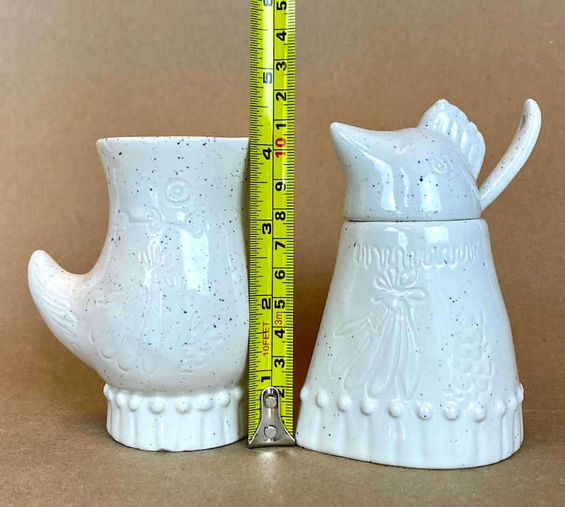 May include: Two white ceramic kitchenware items with a speckled glaze. The item on the left is a pitcher shaped like a boot with a white floral design. The item on the right is a sugar dispenser shaped like a bird with a white floral design.