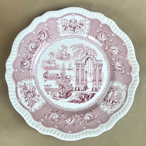 May include: A white ceramic dinner plate with a pink and white floral design. The plate features a central design of a garden scene with an archway, trees, and a boat. The border of the plate is decorated with a repeating pattern of flowers and leaves.