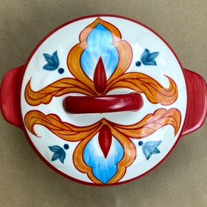 May include: A red and white ceramic bowl with a floral design. The bowl has a red handle and a white interior. The floral design features blue, orange, and red colors.