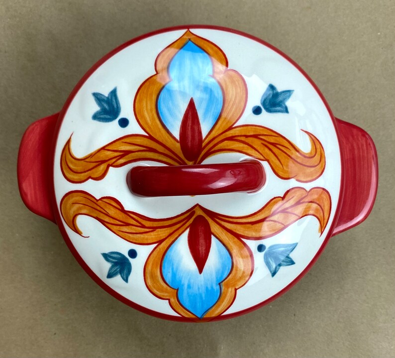 May include: A red and white ceramic bowl with a floral design. The bowl has a red handle and a white interior. The floral design features blue, orange, and red colors.