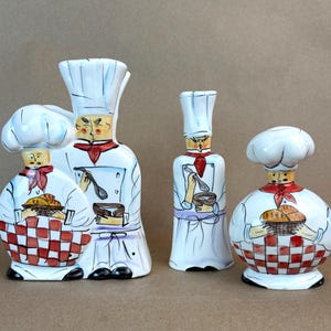 May include: A set of three ceramic kitchen accessories featuring hand-painted chef figures. The pieces include a large double-chef holder, a tall slender chef bottle, and a round chef shaker, all wearing white hats, red neckerchiefs, and checkered aprons.