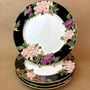 May include: Set of decorative plates with a black border and floral design. The plates feature pink and purple peonies with green leaves and gold accents. The plates have a white center and a gold rim.