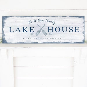 May include: White wooden sign with blue text and a blue watercolor background. The sign reads "The Wilson Family Lake House Est. 2020 Lake Tahoe, California". Two crossed oars are depicted in the center of the sign.