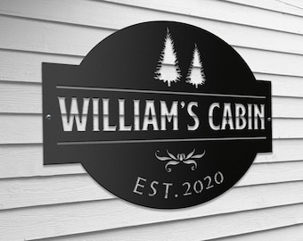 May include: Black metal sign with a rounded top and a white silhouette of two pine trees. The sign reads "William's Cabin. Est. 2020".