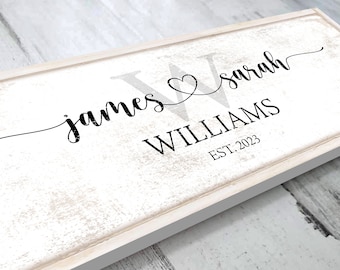 May include: White wooden sign with black text that reads "james & sarah WILLIAMS EST. 2023". The names are written in cursive script with a heart symbol between them.