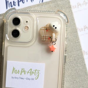 Könnte beinhalten: Eine transparente Handyhülle mit einem weißen und rosafarbenen Anhänger. Der Anhänger ist eine kleine Wolke mit einem karierten Muster und einem rosa Stern, der daran hängt. Die Handyhülle hat einen Aufkleber mit der Aufschrift "Mo Po Artz by Amy Trieu - Etsy UK".