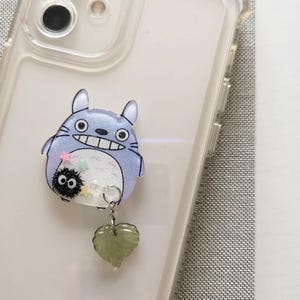 May include: A clear phone case with a blue and white Totoro character charm attached. The charm has a small black and white soot sprite character attached to it. A green heart-shaped charm hangs from the Totoro charm.