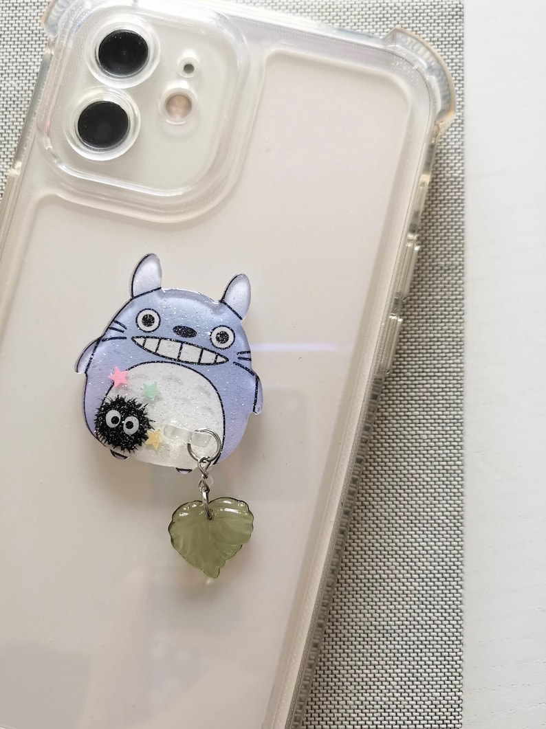 May include: A clear phone case with a blue and white Totoro character charm attached. The charm has a small black and white soot sprite character attached to it. A green heart-shaped charm hangs from the Totoro charm.