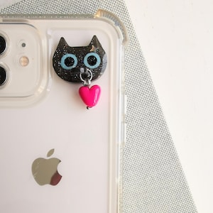 May include: A black cat-shaped phone charm with blue eyes and a pink heart pendant. The charm is attached to a clear phone case. The phone case has a silver apple logo on the back.