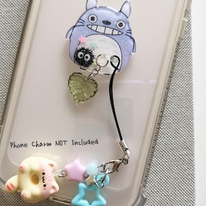 May include: A clear phone case with a Totoro character printed on the back. A black phone strap with a charm attached to it is hanging from the case. The charm features a small black and white character, a green leaf, and a silver ring. The text "Phone Charm NOT Included" is printed on the case.