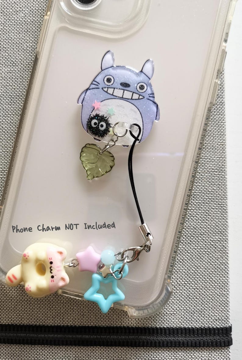 May include: A clear phone case with a Totoro character printed on the back. A black phone strap with a charm attached to it is hanging from the case. The charm features a small black and white character, a green leaf, and a silver ring. The text "Phone Charm NOT Included" is printed on the case.