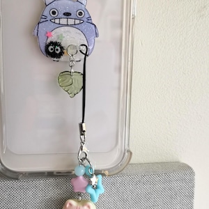 May include: A clear phone case with a blue and white Totoro character charm attached to a black phone strap. The strap has a green leaf charm and a small white cat charm with pink accents.