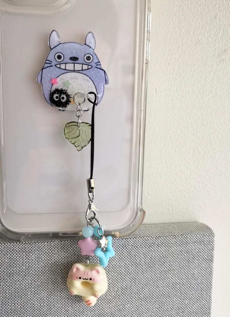 May include: A clear phone case with a blue and white Totoro character charm attached to a black phone strap. The strap has a green leaf charm and a small white cat charm with pink accents.